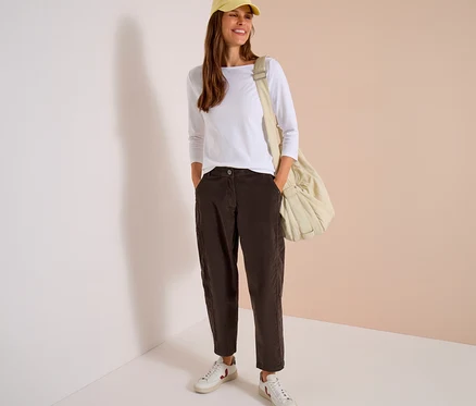 Lächelnde Frau mit gelber Kappe, weißem Langarmshirt und dunkelbrauner CECIL Cordhose, Hände in den Taschen, dazu weiße Sneaker und beiger Schultertasche.