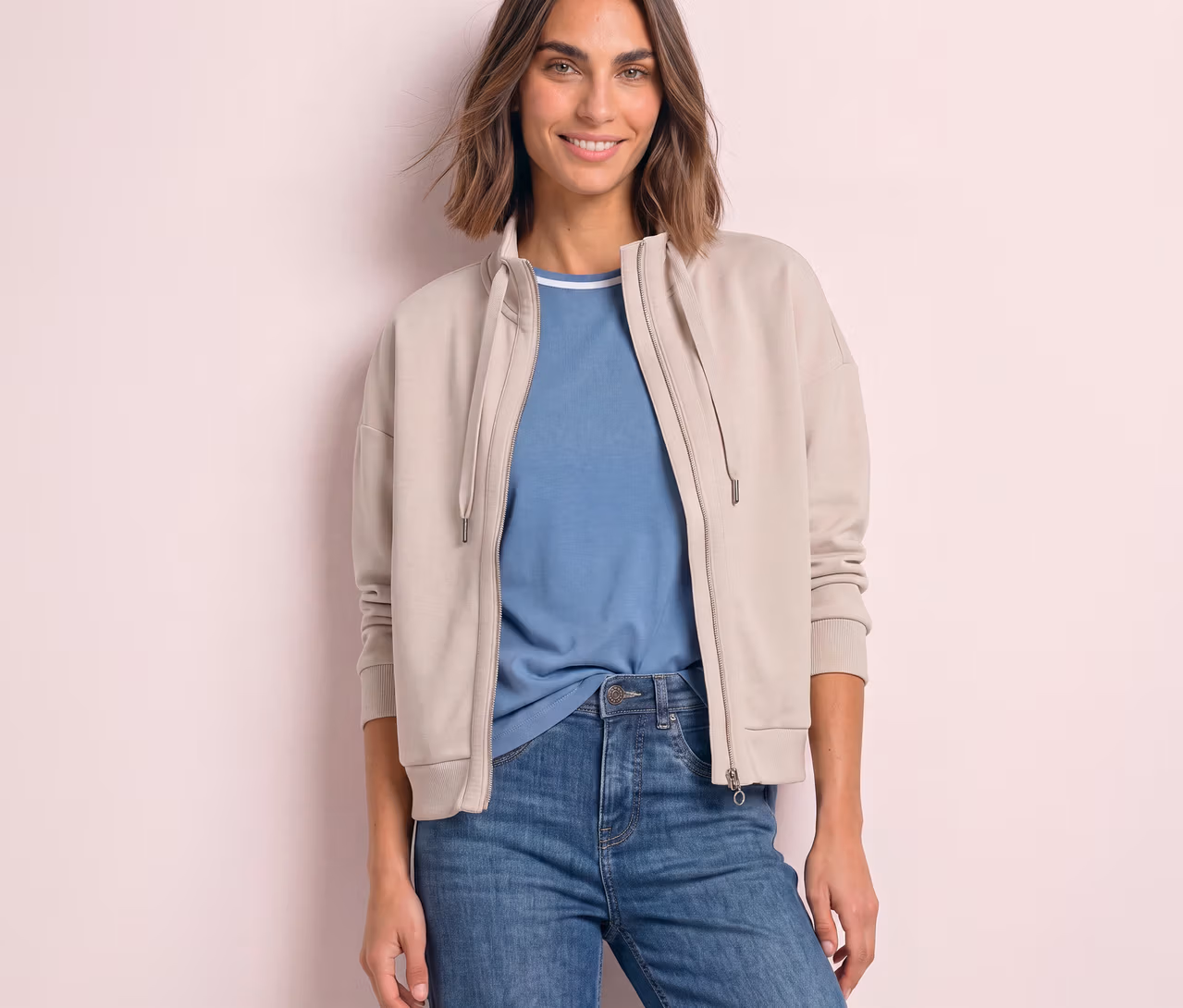 Lächelnde Frau in einer beigen, offenen Street One Sweatjacke über einem blauen T-Shirt und Jeans.