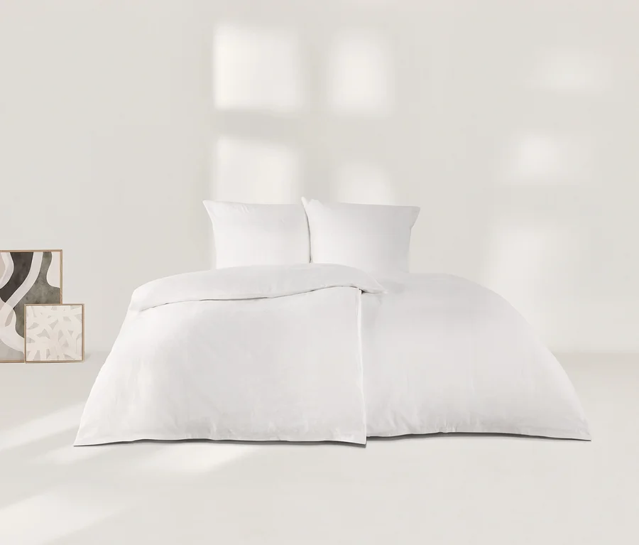 Weiße Leinen-Bettwäsche mit zwei Kissen und einem Bettbezug auf einem Bett in einem minimalistischen weißen Raum. Links lehnen zwei gerahmte abstrakte Kunstdrucke an der Wand.