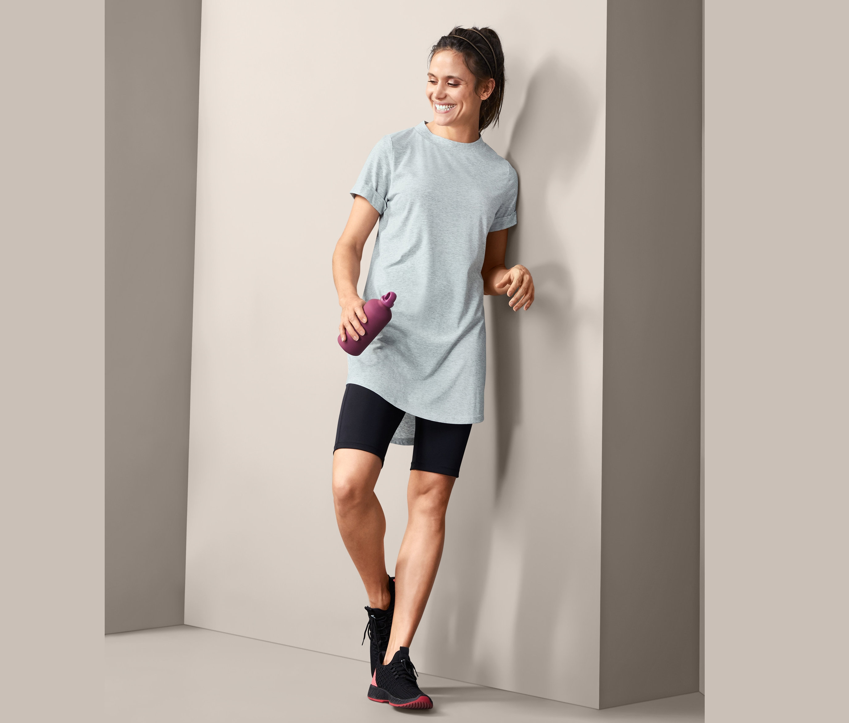 Lächelnde Frau lehnt an einer Wand, hält eine violette Wasserflasche und trägt ein graues Longshirt.