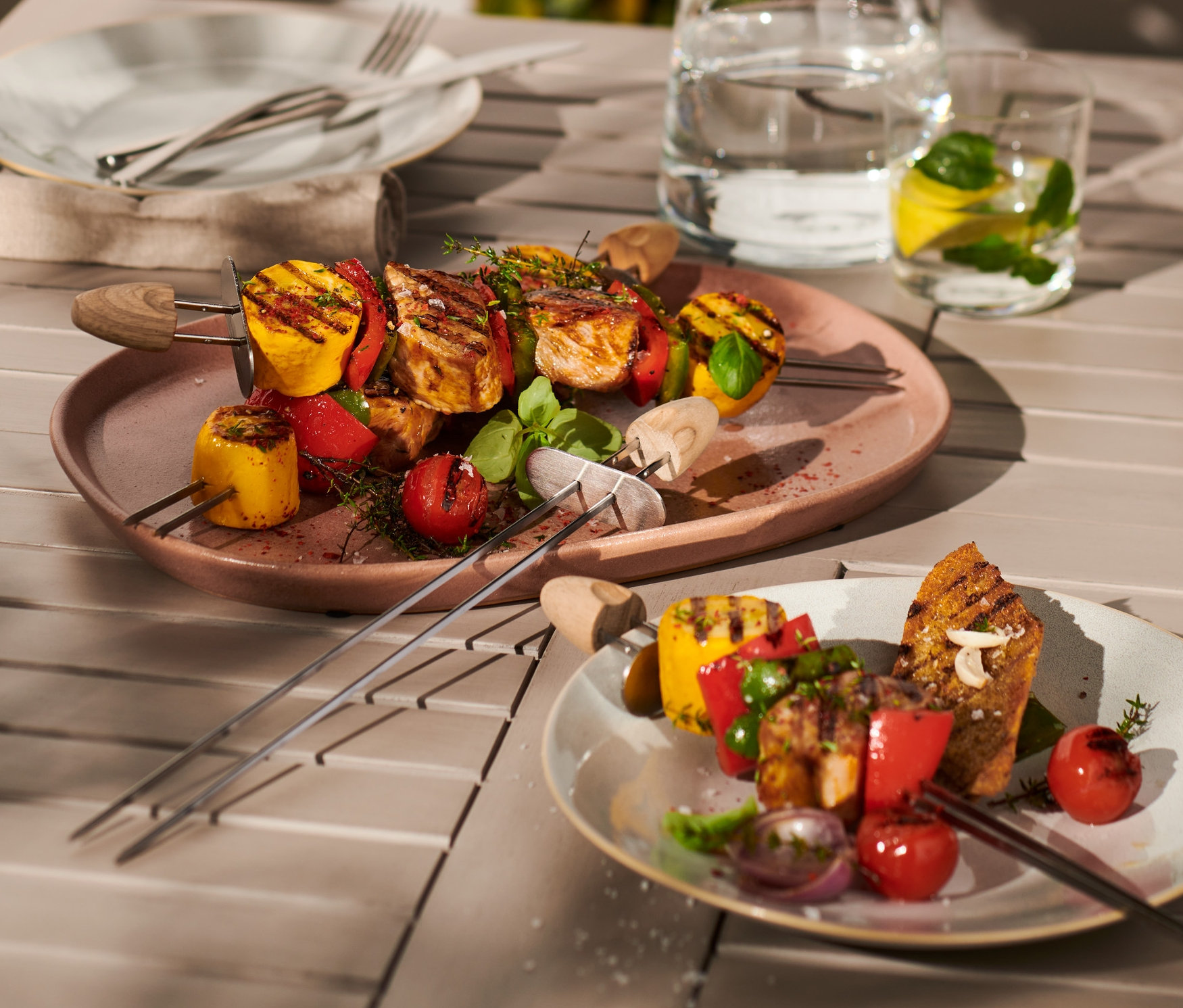 Zwei Teller mit je 4 Schaschlikspießen mit gegrilltem Gemüse und Fleisch auf einem Holztisch.