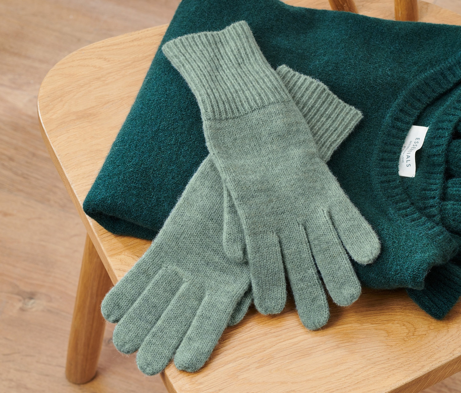 Auf einem Holzstuhl liegen ein grüner Pullover und Strickhandschuhe mit Wolle in Salbeigrün.