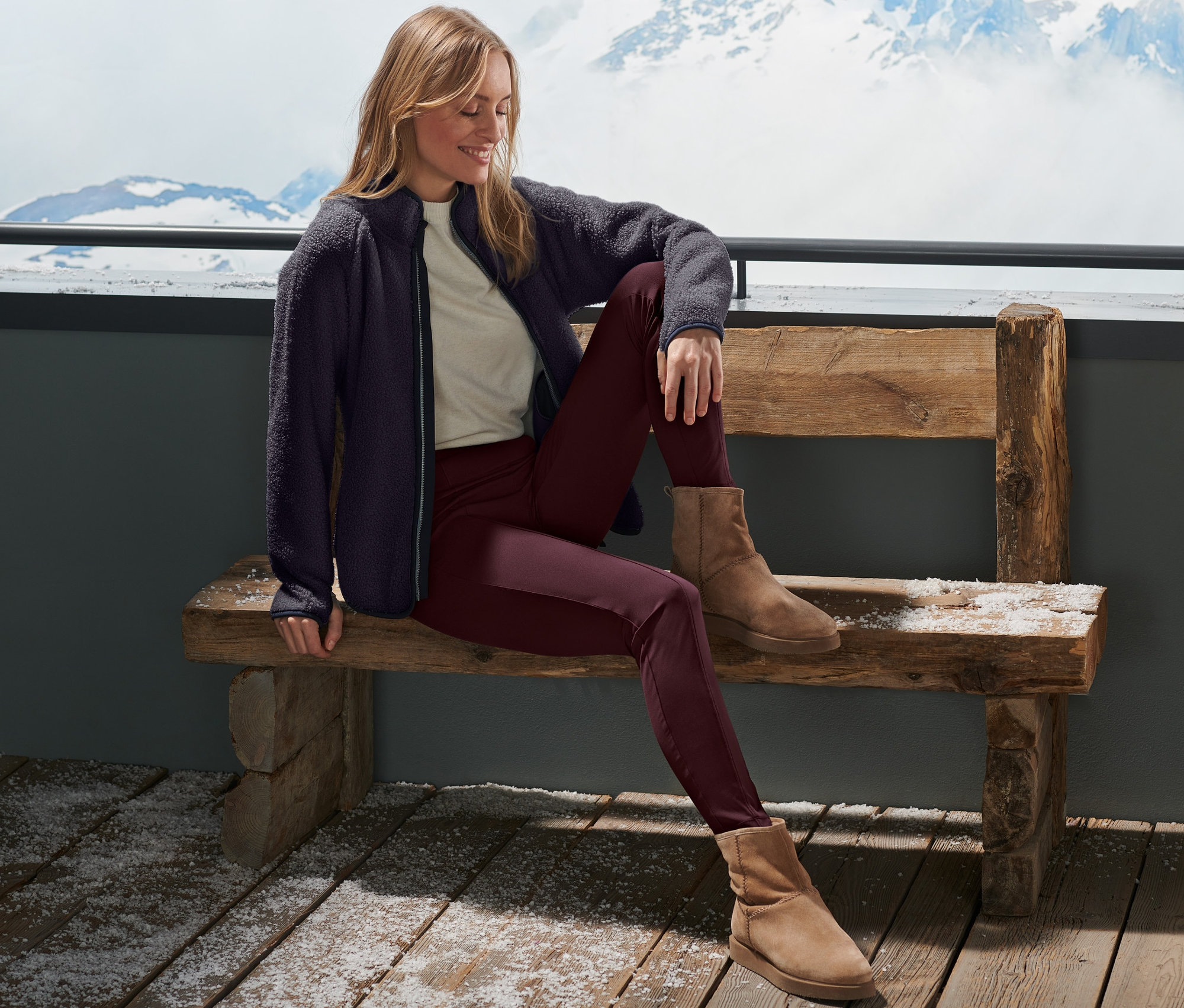Eine Frau sitzt auf einer Bank vor verschneiten Bergen und trägt eine rote Thermotight.