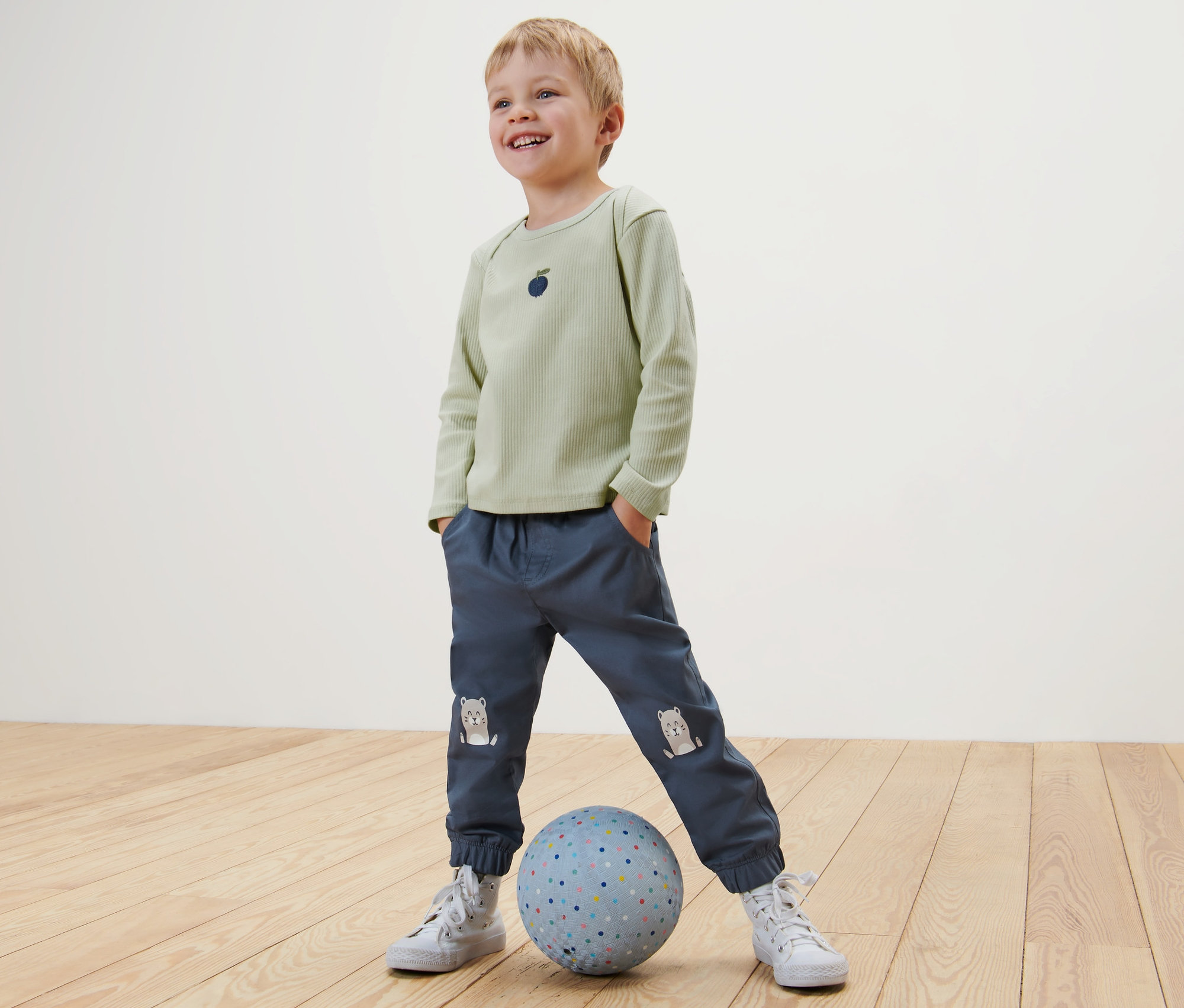 Ein Junge steht mit den Händen in den Hosentaschen und einem Fuß auf einem Ball. Er trägt ein hellgrünes Shirt und blaue Kinder-Pull-on-Pants – Fit »Ella«, Otter.