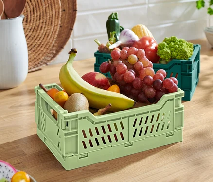 Eine grüne klappbare Ordnungskiste mit Banane, Apfel, Kiwi und Trauben. Im Hintergrund eine grüne klappbare Ordnungskiste mit Gemüse.