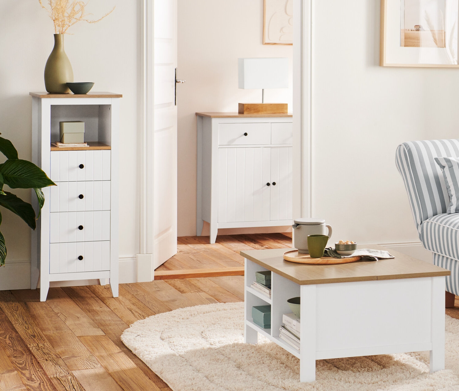 Im Wohnzimmer stehen eine Holz-Stehleuchte »Dreibein« neben einem Sofa und ein Couchtisch mit Klappenfunktion. Im Hintergrund befinden sich eine Kommode mit Schubladen und eine Tischleuchte mit Holzfuß sowie ein Sideboard.