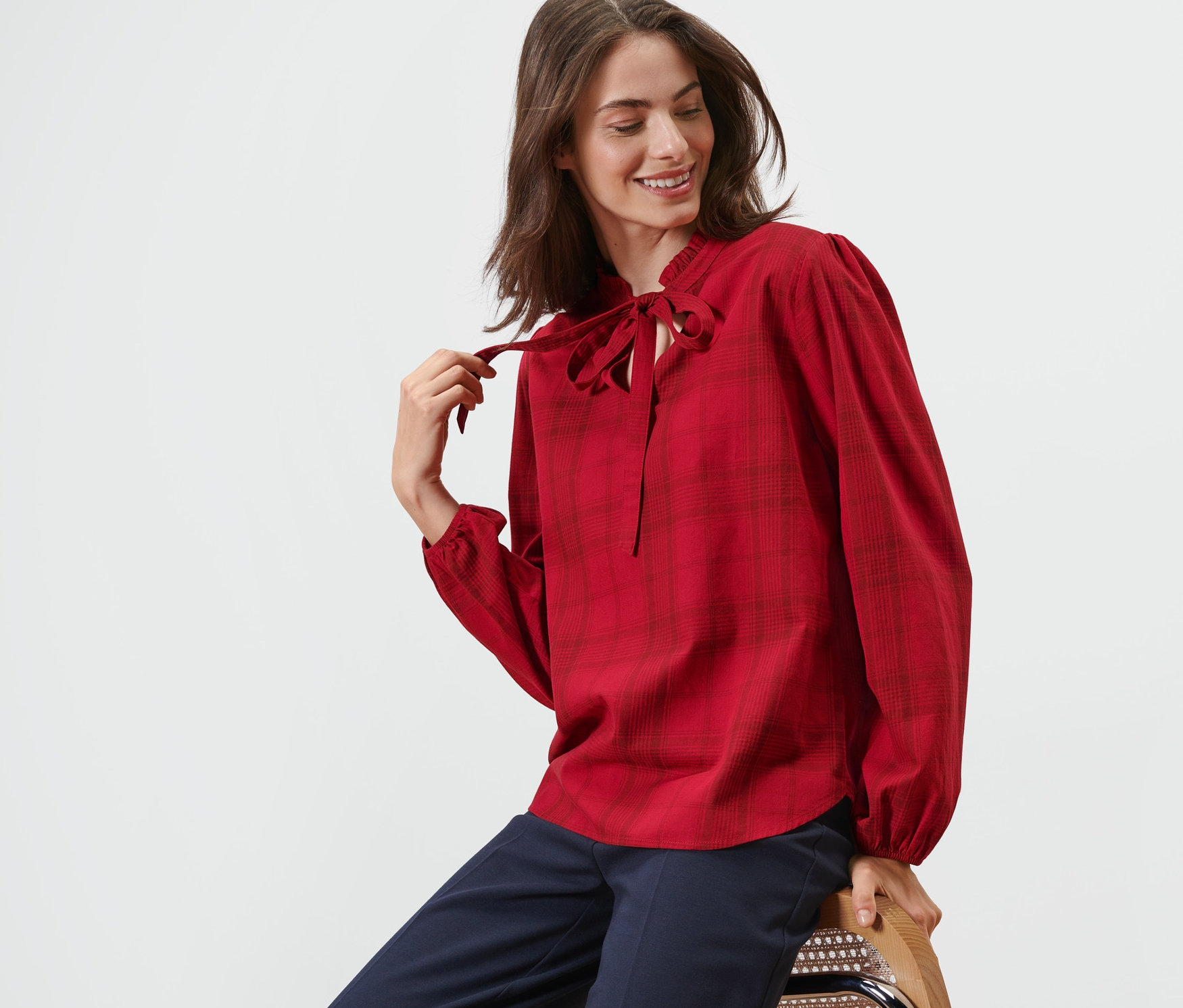 Frau sitzt auf einem Stuhl und trägt eine karierte Schluppenbluse.