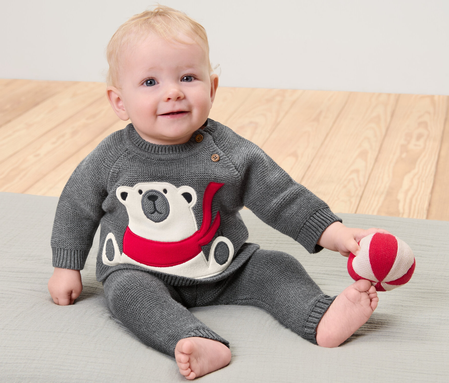 Ein Kind sitzt auf einer grauen Unterlage und spielt mit einem rot-weissen Ball. Es trägt einen grauen Kinder-Strickpullover mit Eisbär-Applikation.
