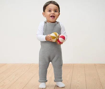 Ein Kind steht und hält zwei Bälle in einer grauen Baby-Merino-Latzhose.