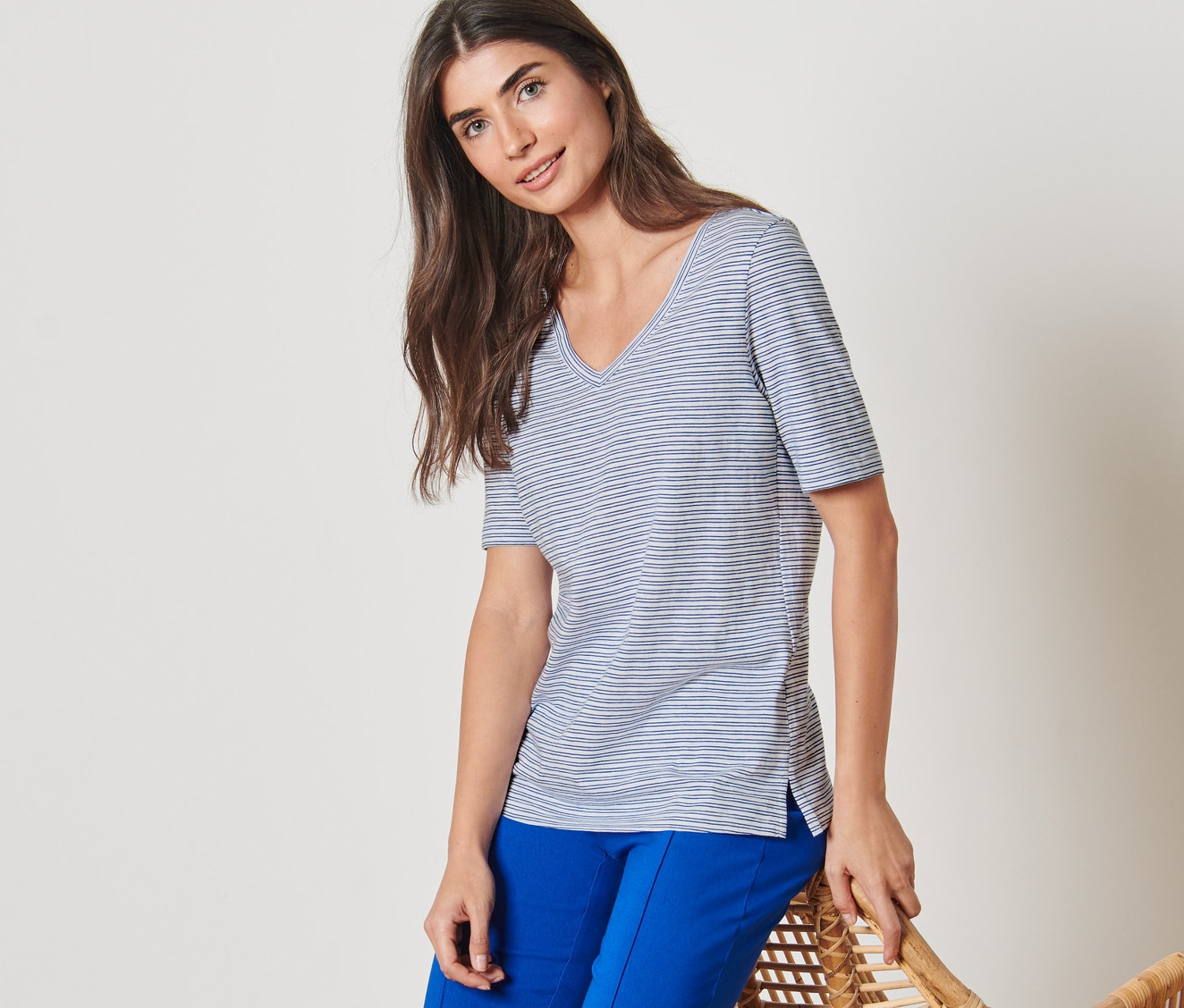 Eine Frau, die sich an einen Rattansessel lehnt, trägt ein gestreiftes Shirt und blaue Hosen.