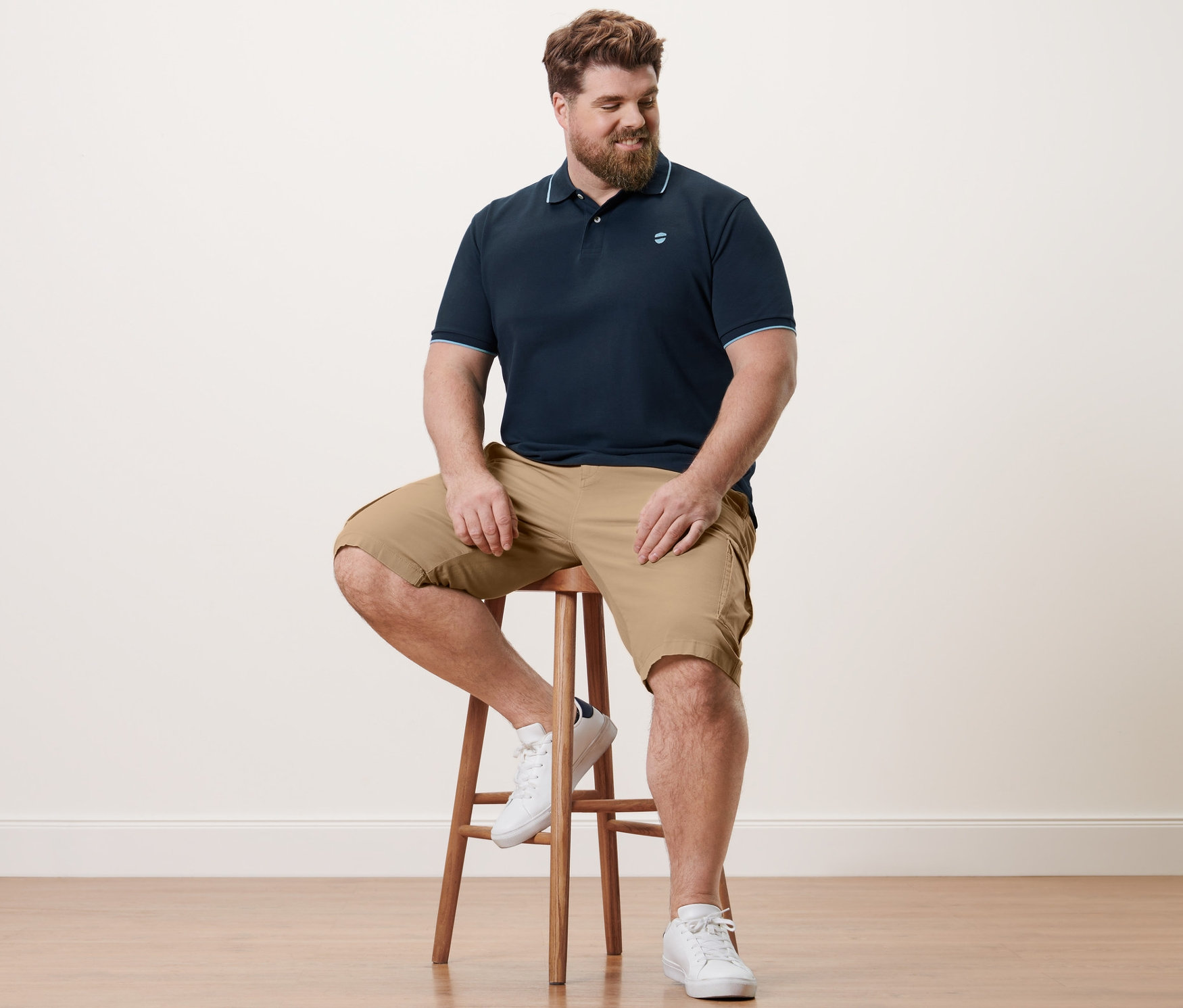 Mann sitzt auf einem Hocker, trägt ein marineblaues Piqué-Poloshirt und sandfarbene Cargo-Shorts.