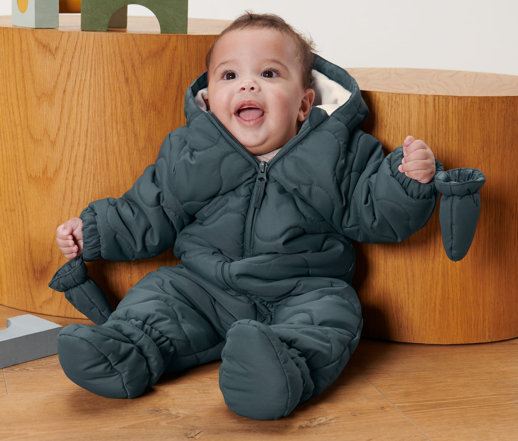 Ein lächelndes Baby sitzt auf dem Boden und trägt einen Baby-Winteroverall.