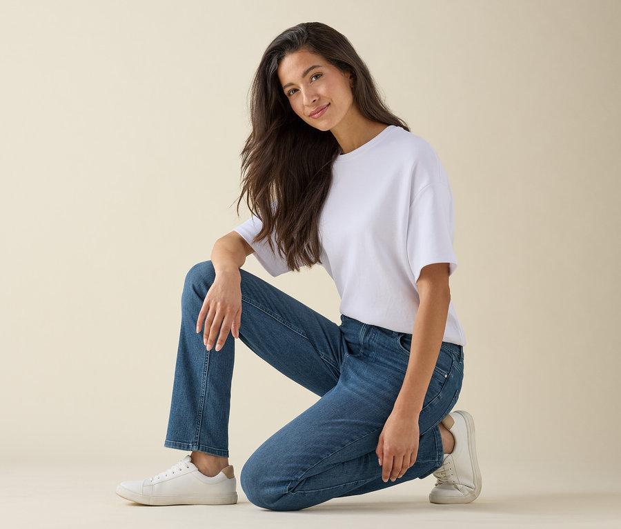 Eine Frau kniet auf einem Knie und lächelt, sie trägt ein weißes T-Shirt, blaue Jeans und weiße Turnschuhe.