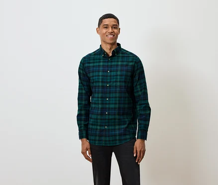Ein kurzhaariger Mann posiert vor einem weißen Hintergrund und trägt ein grün-blau kariertes Langarmhemd und schwarze Jeans.