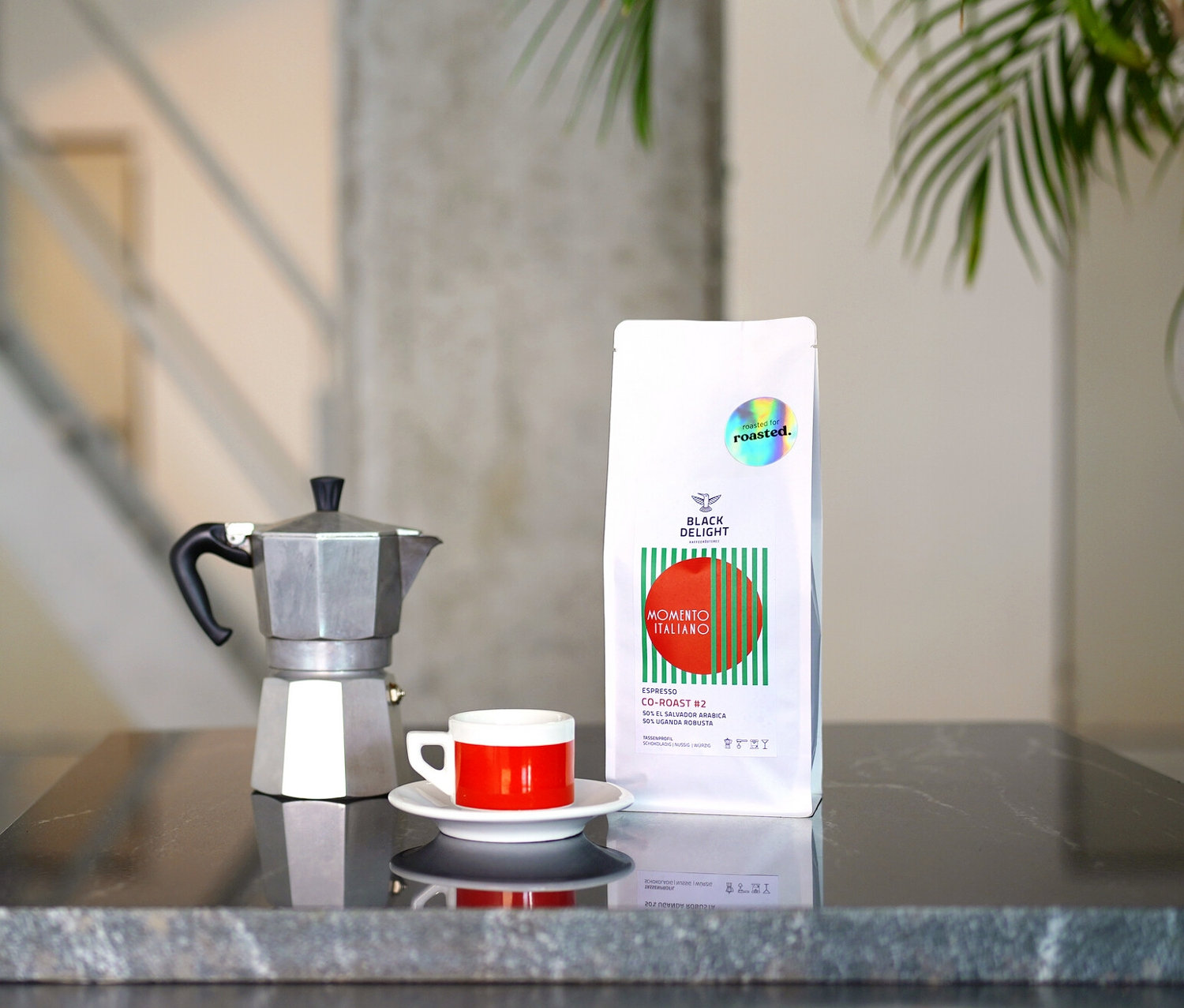 Auf einem Tisch stehen eine Kaffeemaschine, eine Tasse und eine Packung Black Delight x roasted. - Momento Italiano Co-Roast #2 Espresso.