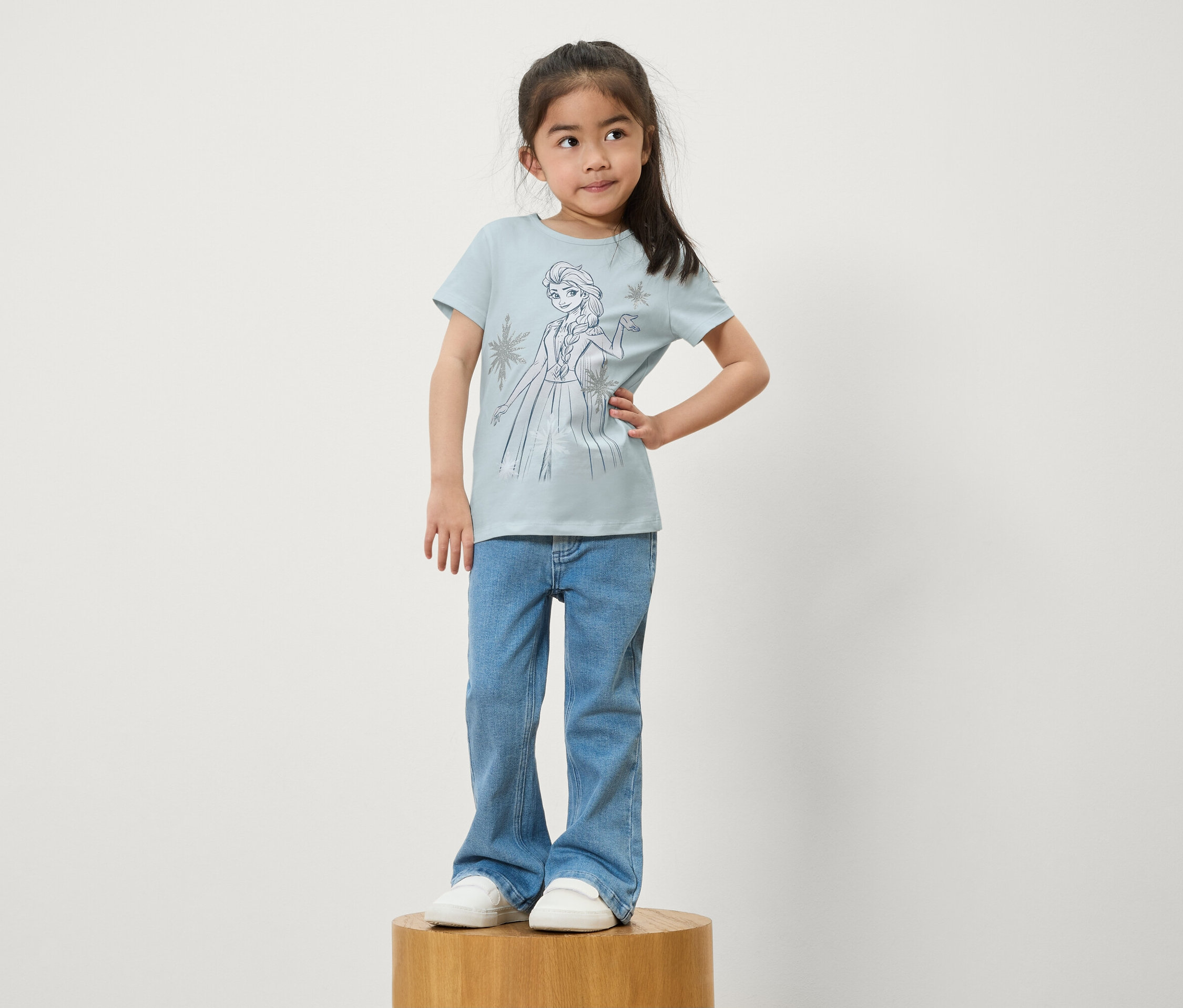 Ein Mädchen steht auf einem Holzsockel. Sie trägt blaue Jeans und ein hellblaues T-Shirt mit einem Aufdruck von Elsa aus Die Eiskönigin.