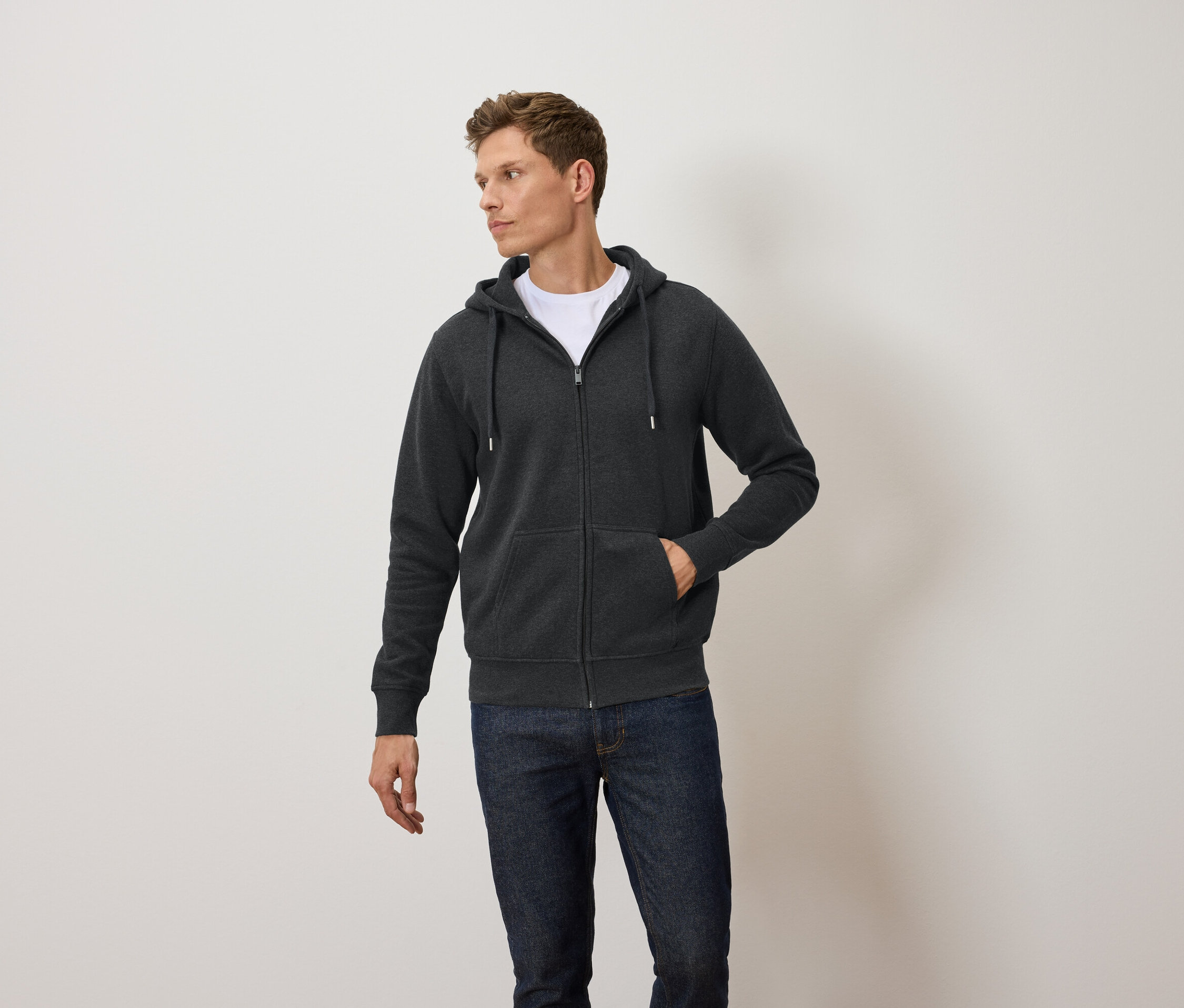 Mann trägt einen dunkelgrauen Kapuzenpullover mit Reißverschluss, ein weißes T-Shirt und blaue Jeans.