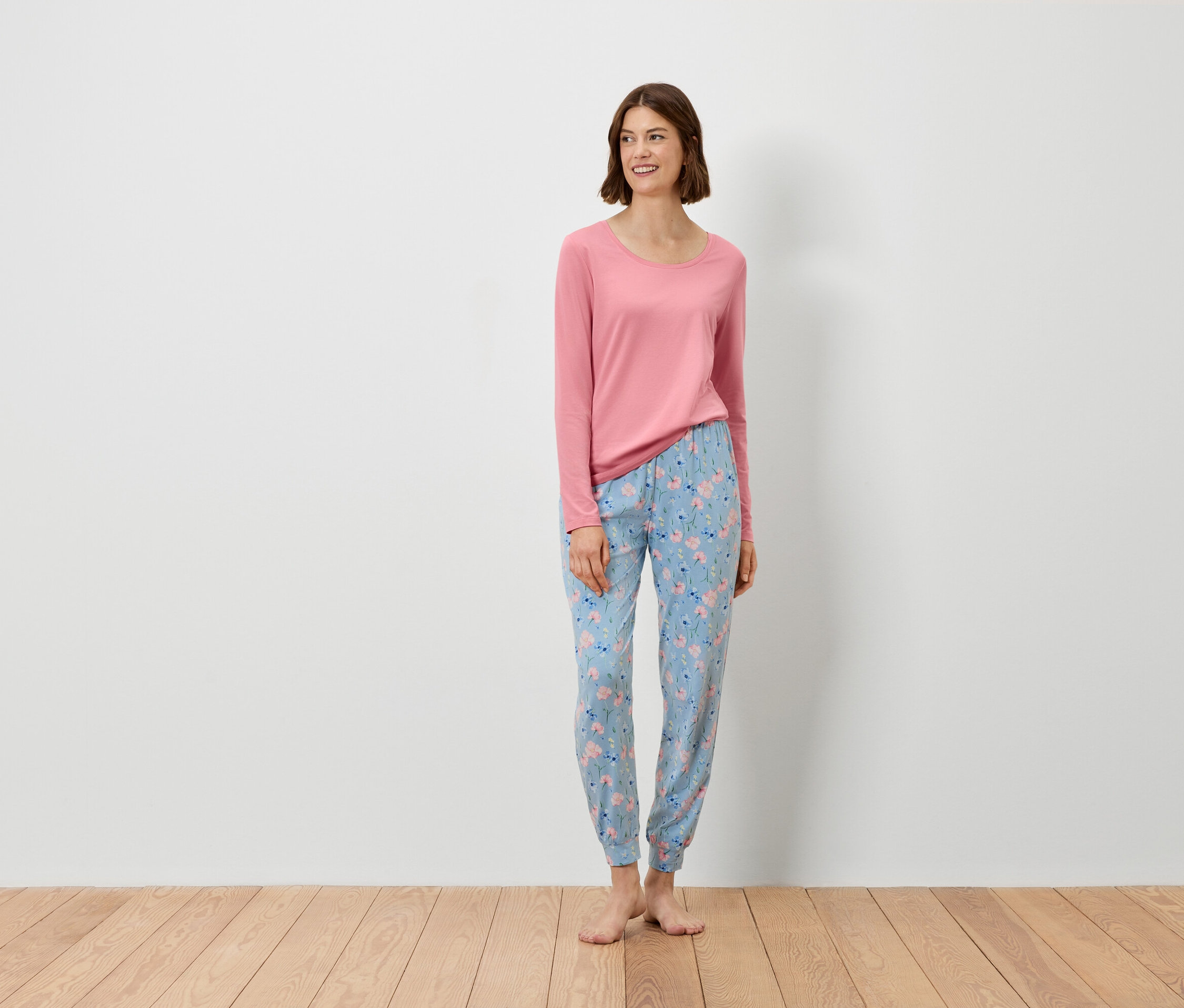 Eine Frau steht und trägt ein rosa Langarmshirt und eine blaue Hose mit Blumenmuster.