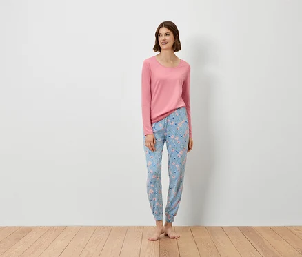Eine Frau steht und trägt ein rosa Langarmshirt und eine blaue Hose mit Blumenmuster.