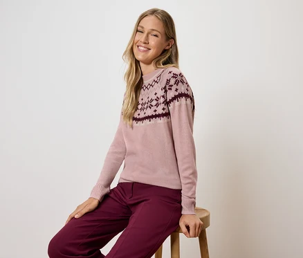 Frau sitzt auf einem Hocker und trägt einen rosa Pullover mit Muster und bordeauxfarbene Hosen.