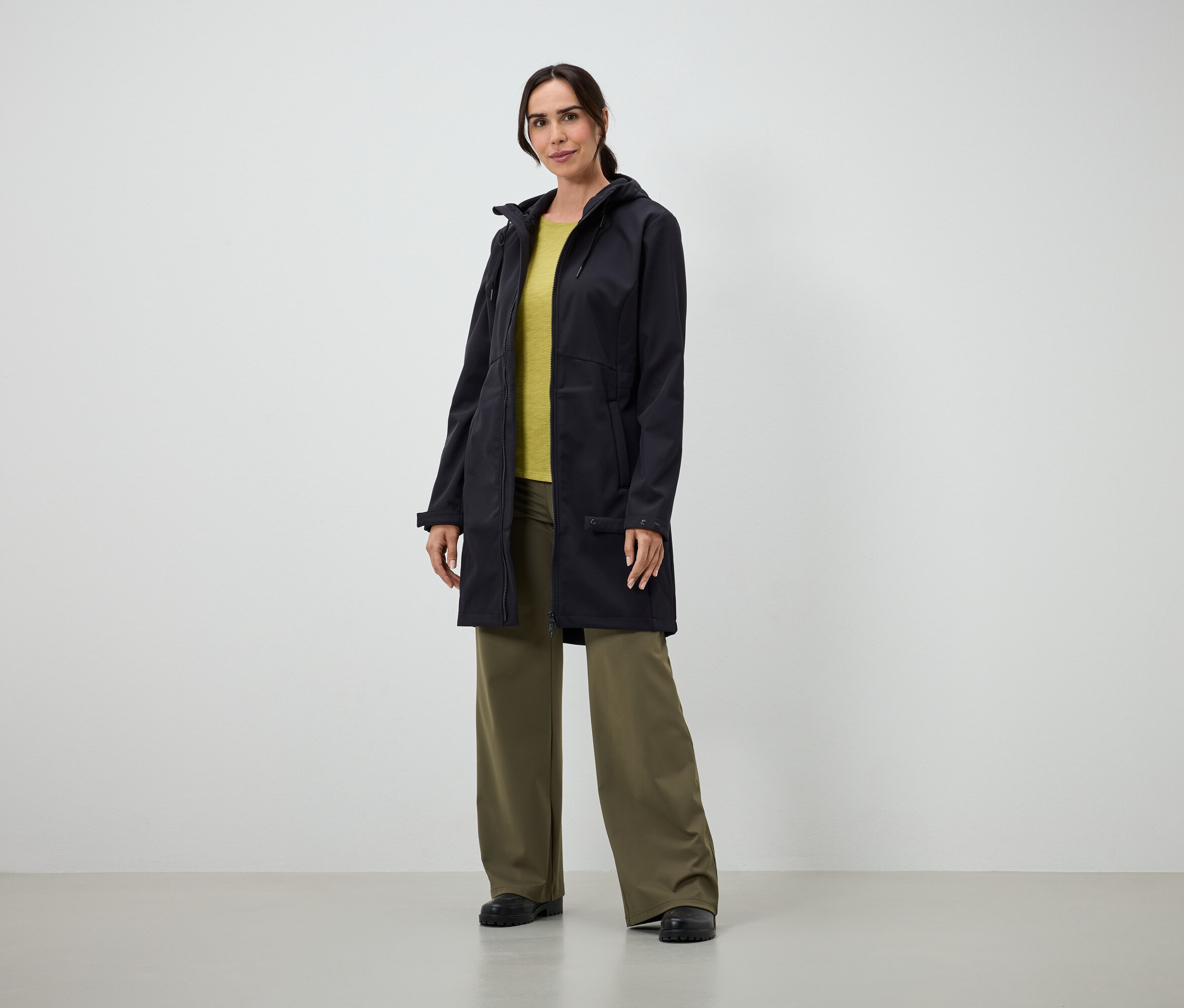 Frau in langem schwarzen Mantel, gelbem T-Shirt und weiter grüner Hose steht drinnen.