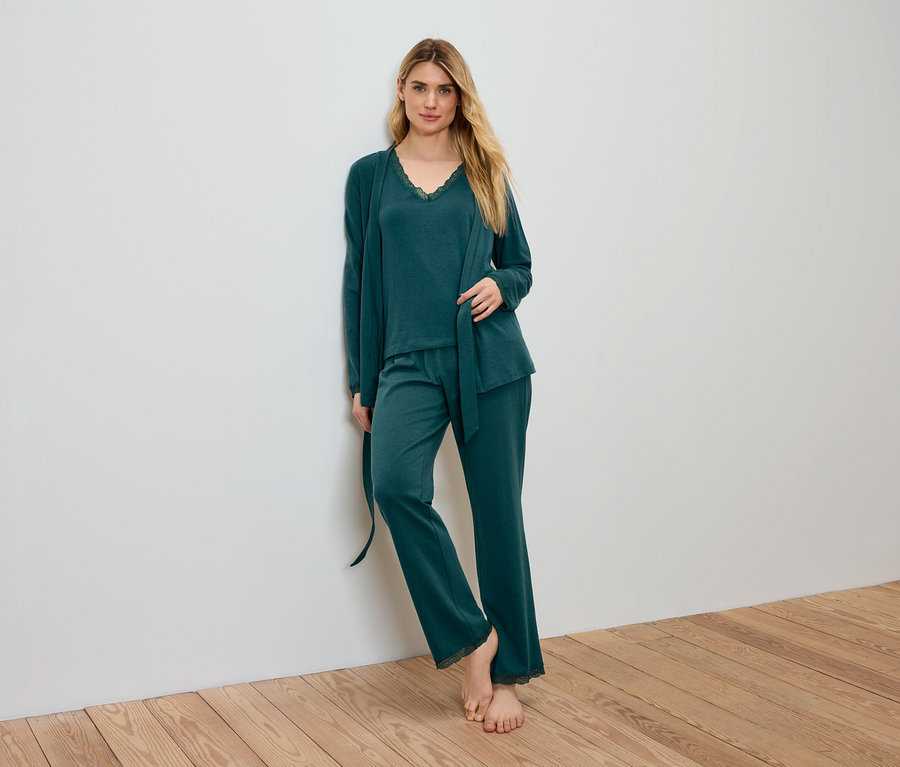 Blonde Frau in grünem Pyjama und Morgenmantel steht an einer Wand.