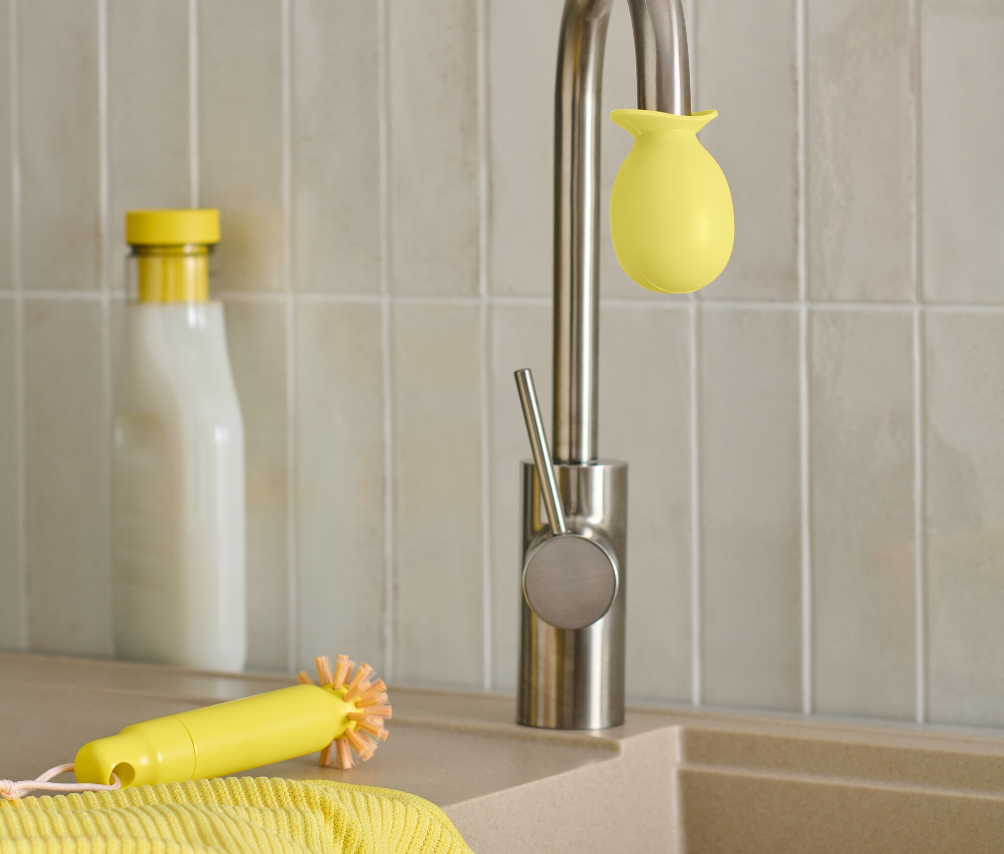 Eine Edelstahlarmatur mit gelbem Schwamm, eine gelbe Bürste und ein Putzlappen liegen auf dem Waschbecken neben einer Flasche.