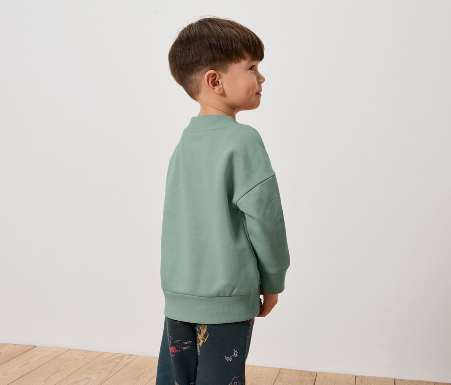 Ein Junge in einem grünen Sweatshirt und gemusterten Hosen steht seitlich.