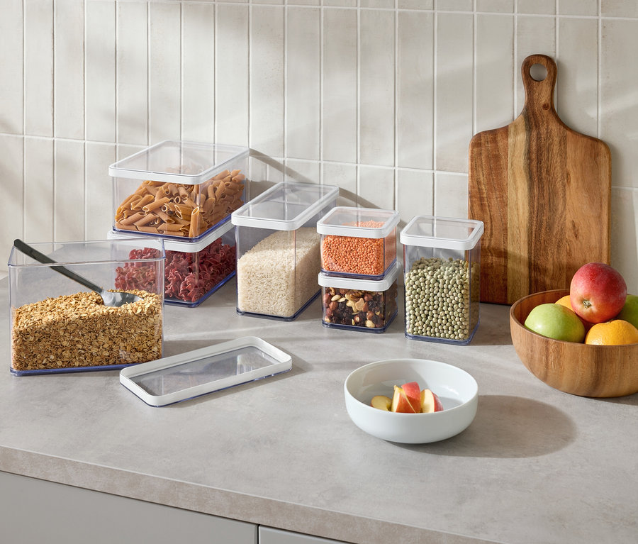 Auf einer Küchentheke stehen mehrere durchsichtige Vorratsbehälter mit verschiedenen Lebensmitteln, ein Holzschneidebrett und eine Schüssel mit Obst.