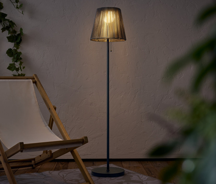 Eine beleuchtete Stehlampe mit Lampenschirm steht neben einem Liegestuhl aus Holz.