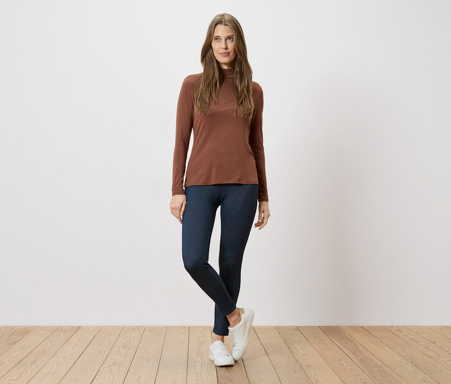 Frau in braunem Rollkragenshirt, blauen Leggings und weißen Turnschuhen steht vor einer weißen Wand auf Holzboden.
