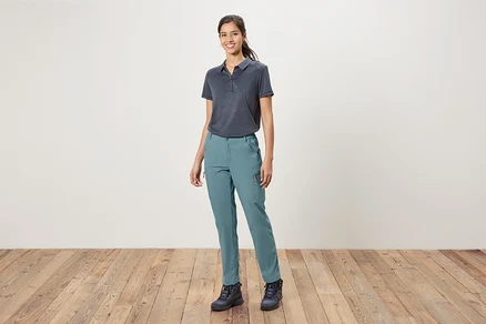 Eine Frau steht vor einer weißen Wand und trägt ein blaues Polohemd, blaue Hosen und Wanderschuhe.