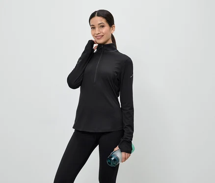 Frau in schwarzem Sportoberteil mit Reißverschluss und Leggings hält eine Wasserflasche vor einem hellen Hintergrund.