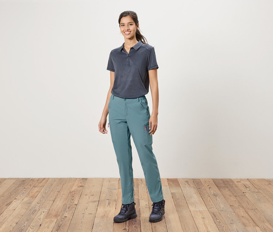 Eine Frau steht vor einer weißen Wand und trägt ein blaues Polohemd, blaue Hosen und Wanderschuhe.