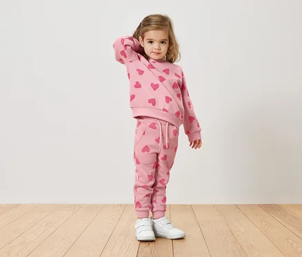 Mädchen mit blonden Haaren trägt einen rosafarbenen Pullover und eine Hose mit Herzmuster und weiße Turnschuhe.