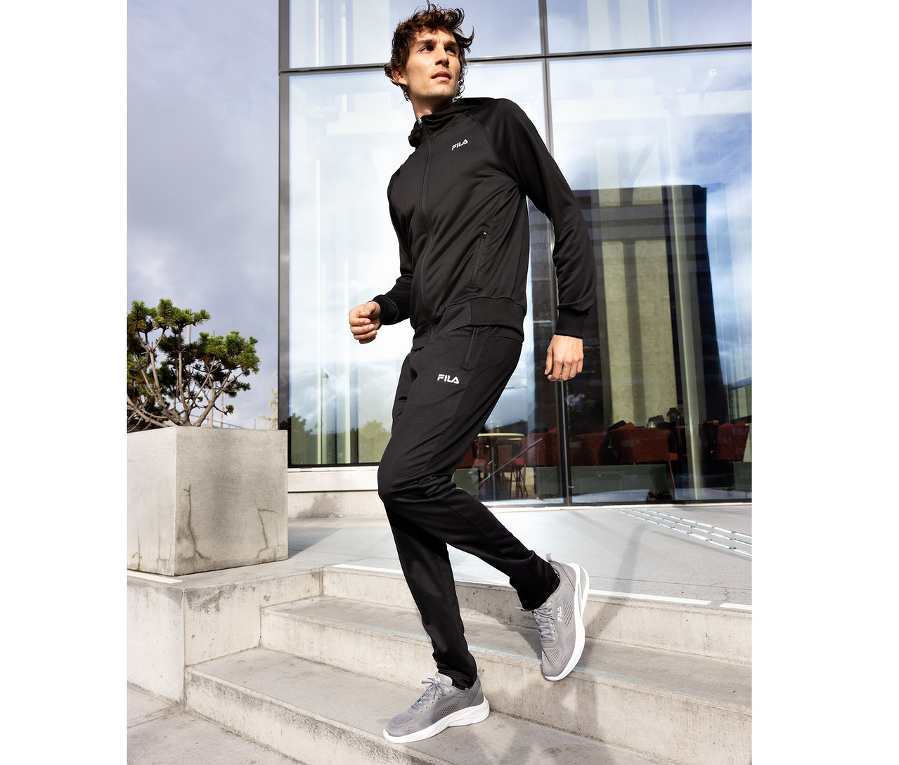 Mann in schwarzem FILA Trainingsanzug mit FILA Herren-Funktions-Sweathose und Kapuzenjacke sowie grauen Sportschuhen, geht eine Betontreppe vor einem modernen Glasgebäude hinunter.