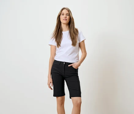 Frau mit langen braunen Haaren, weißem T-Shirt und schwarzen Shorts mit einer Hand in der Tasche vor einem weißen Hintergrund.