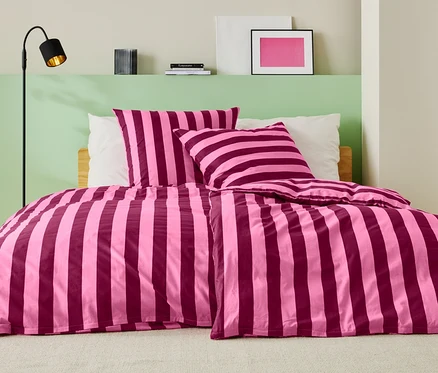Bett mit rosa-weinroter Streifenbettwäsche, zwei Kissen und einer Stehlampe.