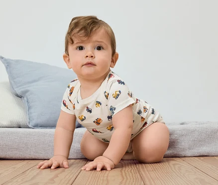 Ein Baby krabbelt auf einem Holzboden, bekleidet mit einem kurzärmeligen Body mit einem Muster aus Baufahrzeugen.