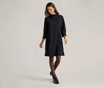 Frau in schwarzem, knielangem Strickkleid mit Rollkragen, dunkler Strumpfhose und schwarzen Schuhen vor hellem Hintergrund.