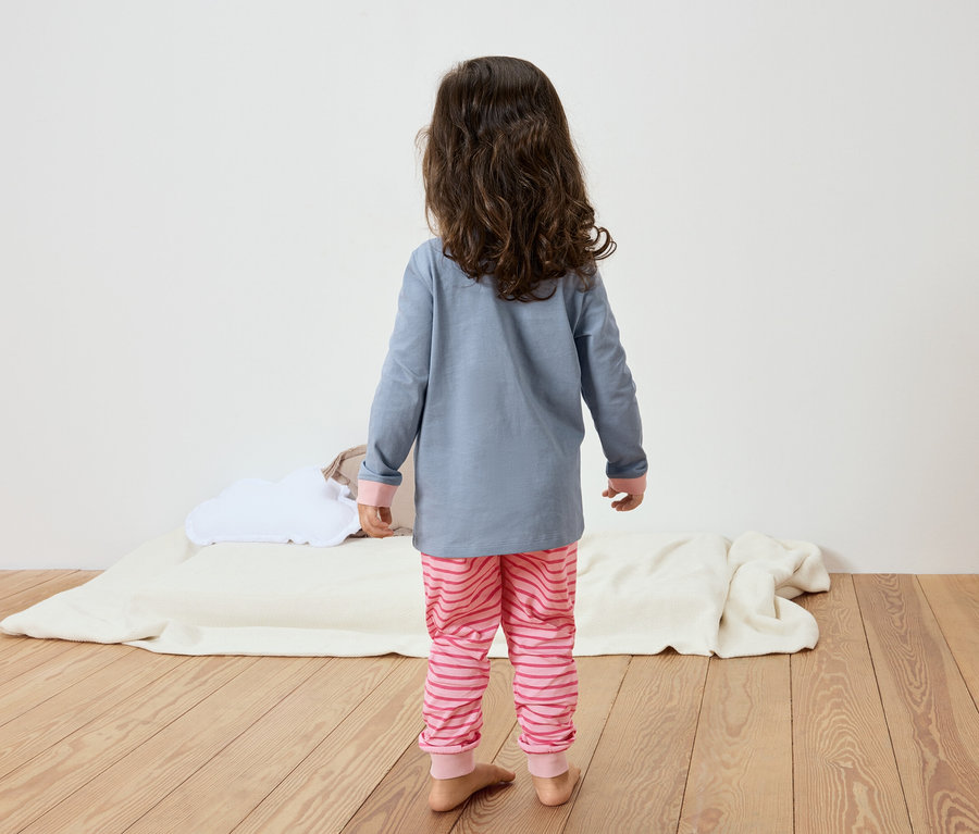 Ein kleines Mädchen mit langen, lockigen, braunen Haaren steht mit dem Rücken zum Betrachter, trägt einen blaugrauen Pyjamaoberteil und eine rosafarben gestreifte Pyjamahose und steht auf einem Holzboden.
