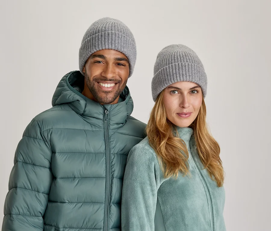 Ein lächelnder Mann und eine Frau tragen graue Strickmützen. Der Mann trägt eine türkisfarbene Steppjacke, die Frau eine helltürkisfarbene Fleecejacke.