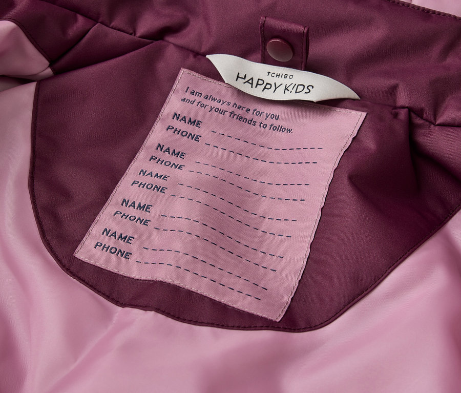 Nahaufnahme eines rosa Etiketts, das in eine lila-rosa Jacke eingenäht ist. Das Etikett trägt oben die Aufschrift "TCHIBO HAPPY KIDS", gefolgt von "I am always here for you and for your friends to follow.". Darunter befinden sich fünf Zeilenpaare für "NAME" und "PHONE".