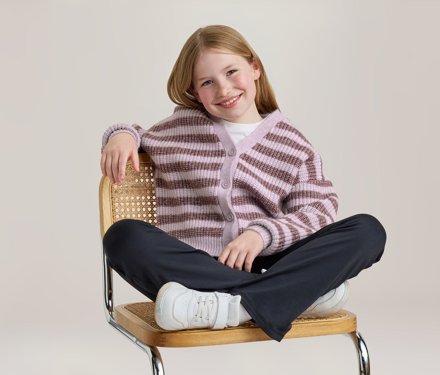 Lächelndes Mädchen sitzt im Schneidersitz auf einem Korbstuhl, trägt eine gestreifte lila-braune Strickjacke, dunkle Hose und weiße Turnschuhe.