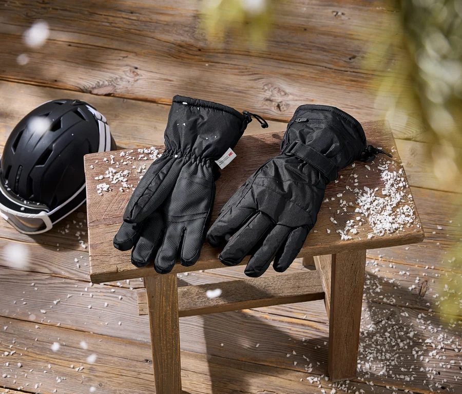 Schwarzer Skihelm mit weißer Skibrille und schwarzen Skihandschuhen auf einem Holzhocker und Holzboden, mit Kunstschnee bestreut.