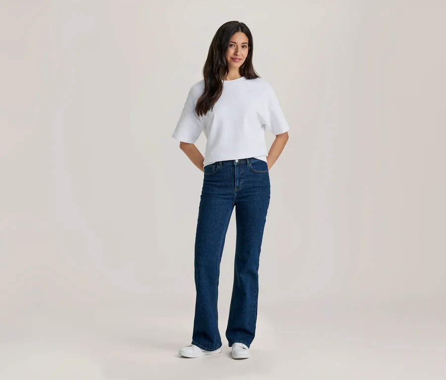 Eine lächelnde Frau mit langen dunklen Haaren trägt ein weißes T-Shirt, dunkelblaue Schlagjeans und weiße Sneaker. Ihre Hände sind hinter dem Rücken.