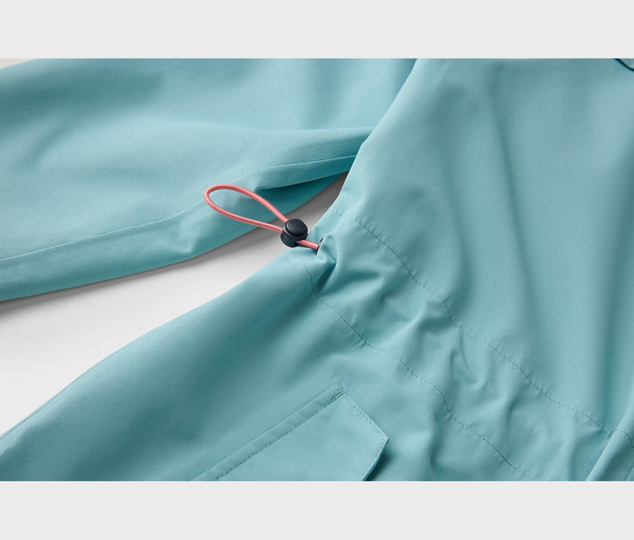Nahaufnahme einer hellblauen Jacke mit einem rosafarbenen Kordelzug und einem schwarzen Stopper, der durch den Stoff läuft. Unten links ist eine Taschenklappe sichtbar.