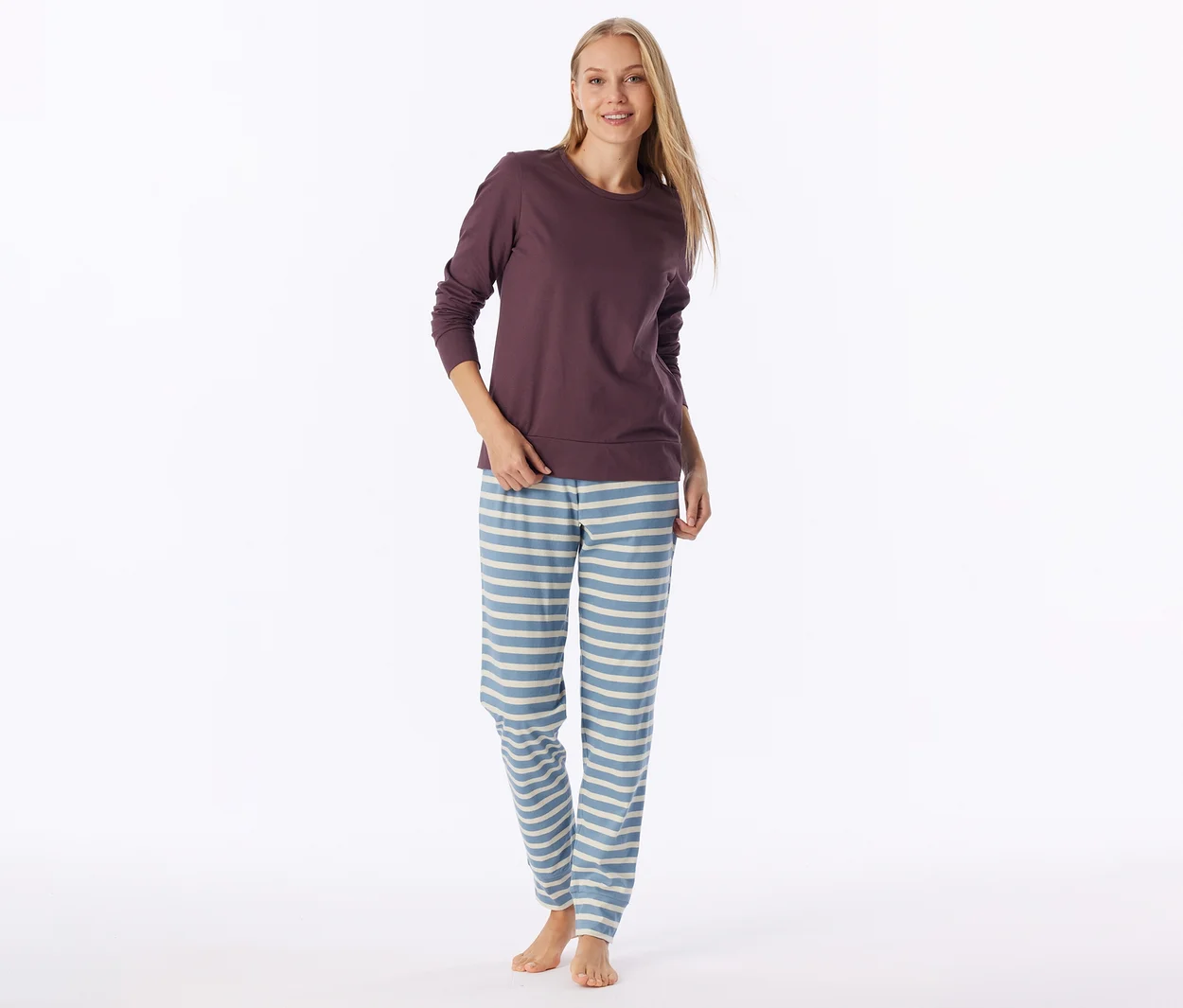 Eine Frau trägt ein dunkelviolettes Langarmshirt und hellblaue Pyjamahosen mit horizontalen cremefarbenen Streifen. Sie hat langes blondes Haar, lächelt und steht barfuß.