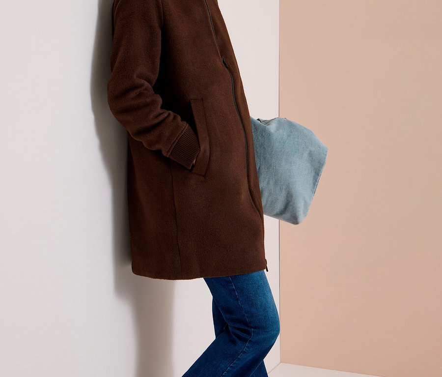 Lächelnde Frau in braunem CECIL Blouson, blauen Jeans und silbernen Sneakers, die an einer Wand lehnt.
