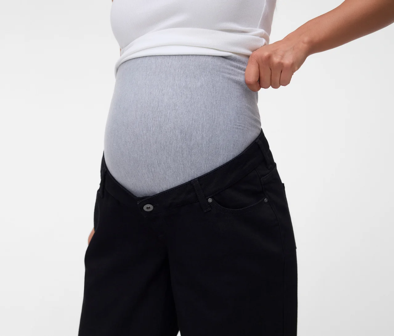 Schwangere Frau in weißem Tanktop und schwarzer Umstandsjeans mit grauem Stretchbund, die ihr Oberteil hochzieht.